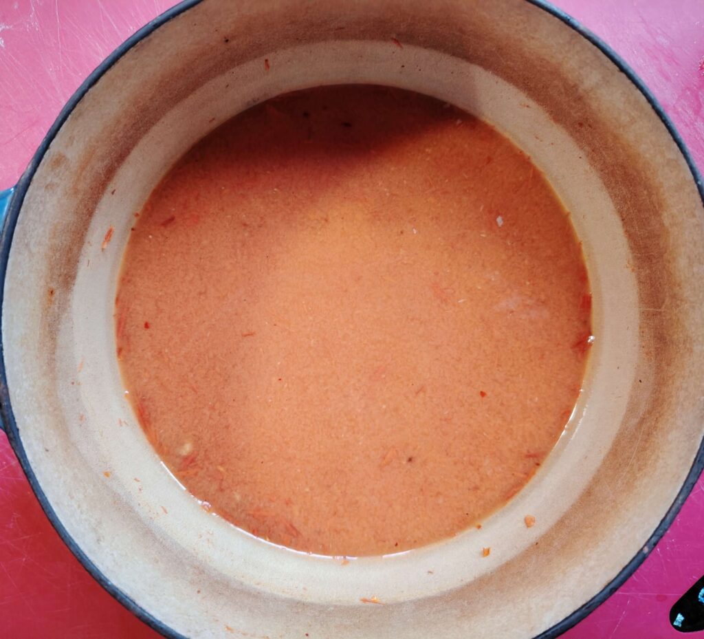 Roasted cherry tomato and carrot sauce simmering in a light enamelled cast iron pot, smooth and reddish-orange with visible seasoning.