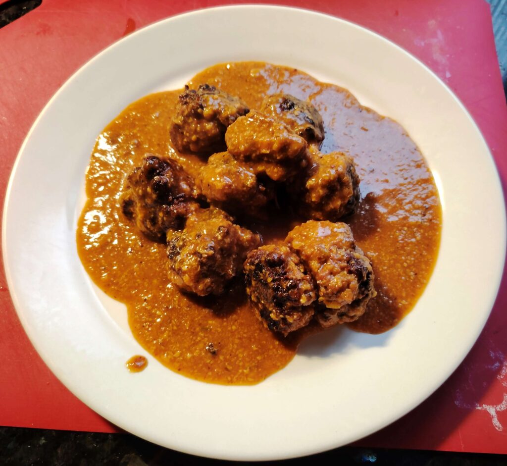 Plate of cooked meatballs covered in a thick, spiced brown-orange sauce, served on a white dish atop a red surface.