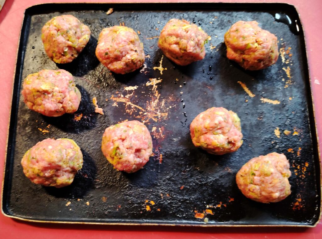 Well used baking tray with nine raw, herb-flecked meatballs evenly spaced, ready for cooking, set against a countertop.