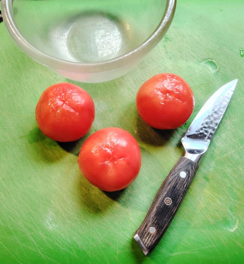 Three peeled tomatoes on a green chopping board with small glass bowl and small knife.