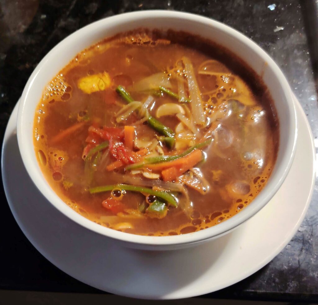 Hearty spicy prawn soup in a white bowl, packed with prawns, vegetables, and noodles in a fiery red broth. A warm, comforting dish with bold colours and bold flavours.