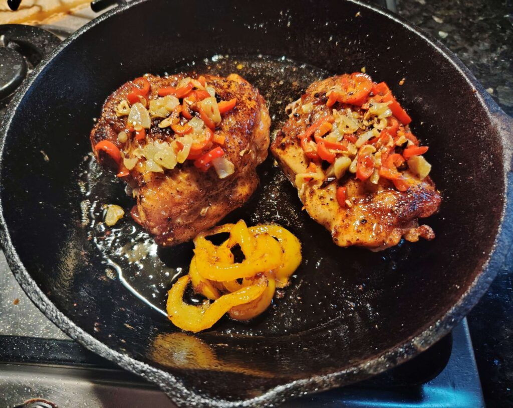 Bone-in chicken thighs cooking in cast iron skillet topped with sautéed peppers and onions.