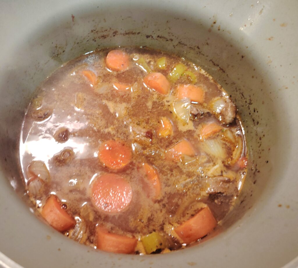A top-down view of a Slow Cooker Spanish Beef Stew simmering in a ceramic pot, showing chunks of beef, carrots, and onions in a rich, dark broth.