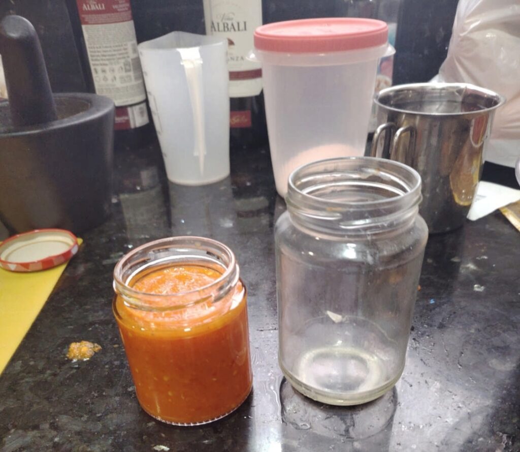 A jar of freshly blended, bright orange fermented habanero hot sauce next to an empty glass jar on a dark kitchen counter.