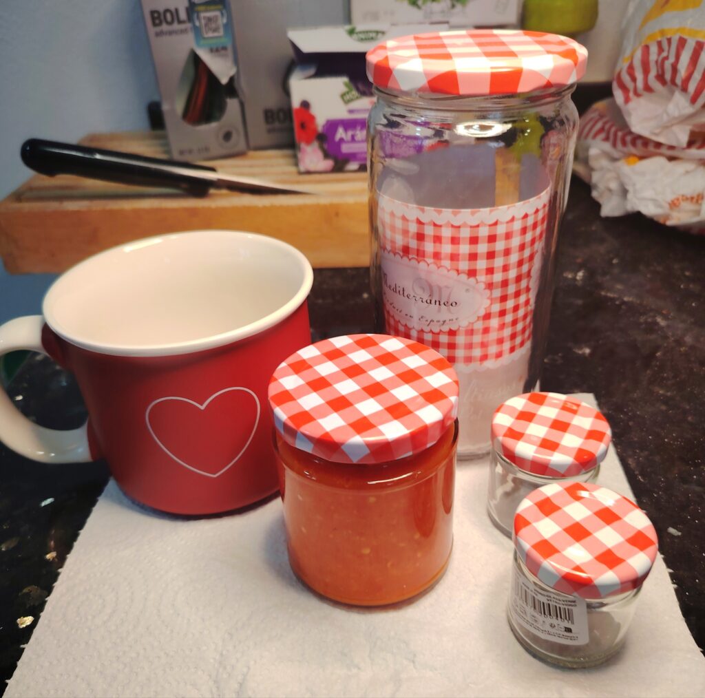 A group of small jars filled with bright orange fermented hot sauce, each with a red‑and‑white chequered lid, arranged on a kitchen counter beside a red mug..