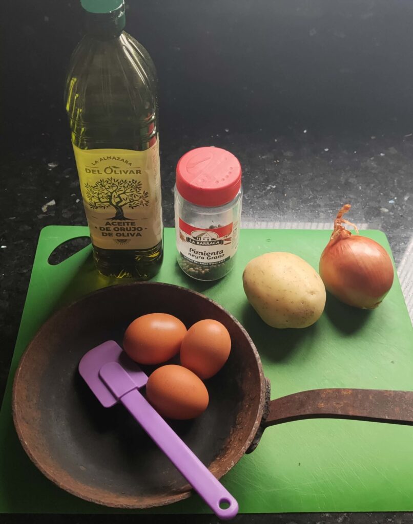 A cutting board with ingredients for cooking, including a potato, an onion, a bottle of olive oil, a container of black pepper, and a frying pan holding three eggs and a spatula.