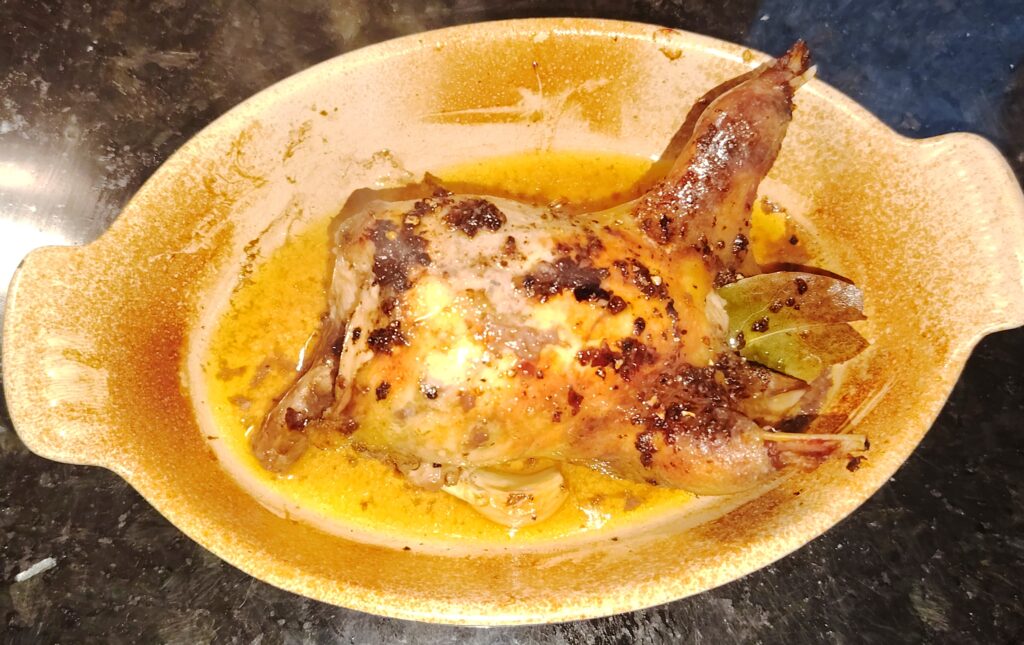 Butter-roasted wild partridge in a ceramic roasting dish, golden and glistening with pan juices and bay leaves