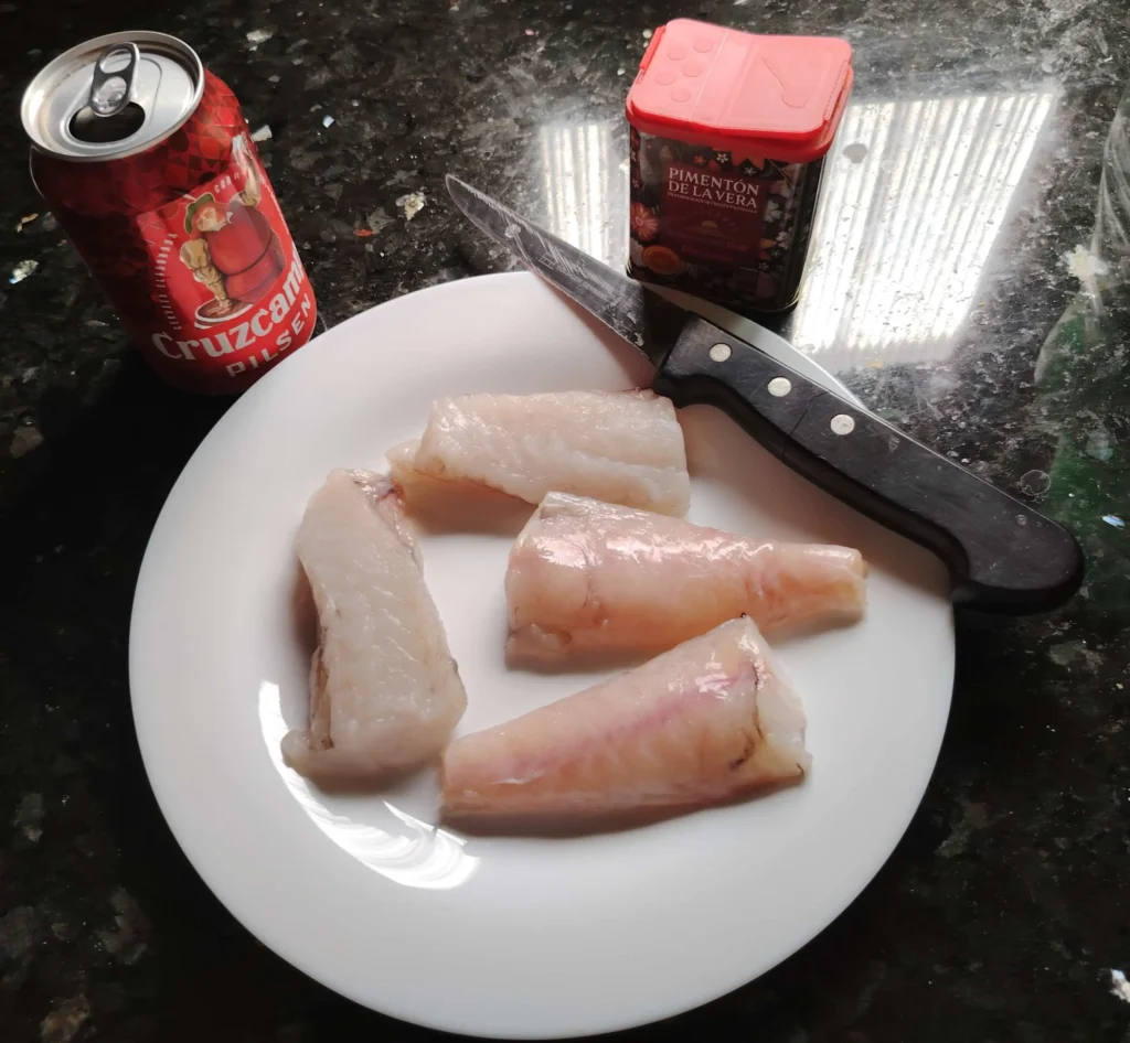 Four skinned monkfish pieces on a white plate, ready for seasoning, with pimentón picante and a knife alongside