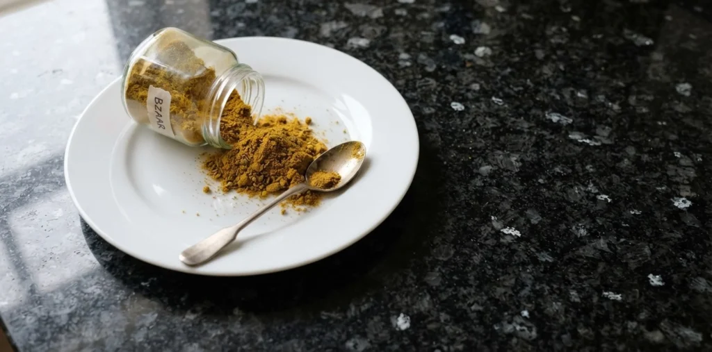 Bzaar spice blend — golden-yellow Libyan spice powder spilling from a labelled glass jar onto a white plate with a silver spoon on a black speckled granite worktop