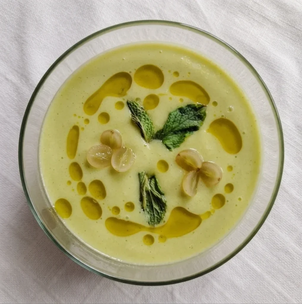 Cucumber avocado gazpacho in a glass bowl with aceite verde, white grapes and fresh mint, overhead shot on white linen