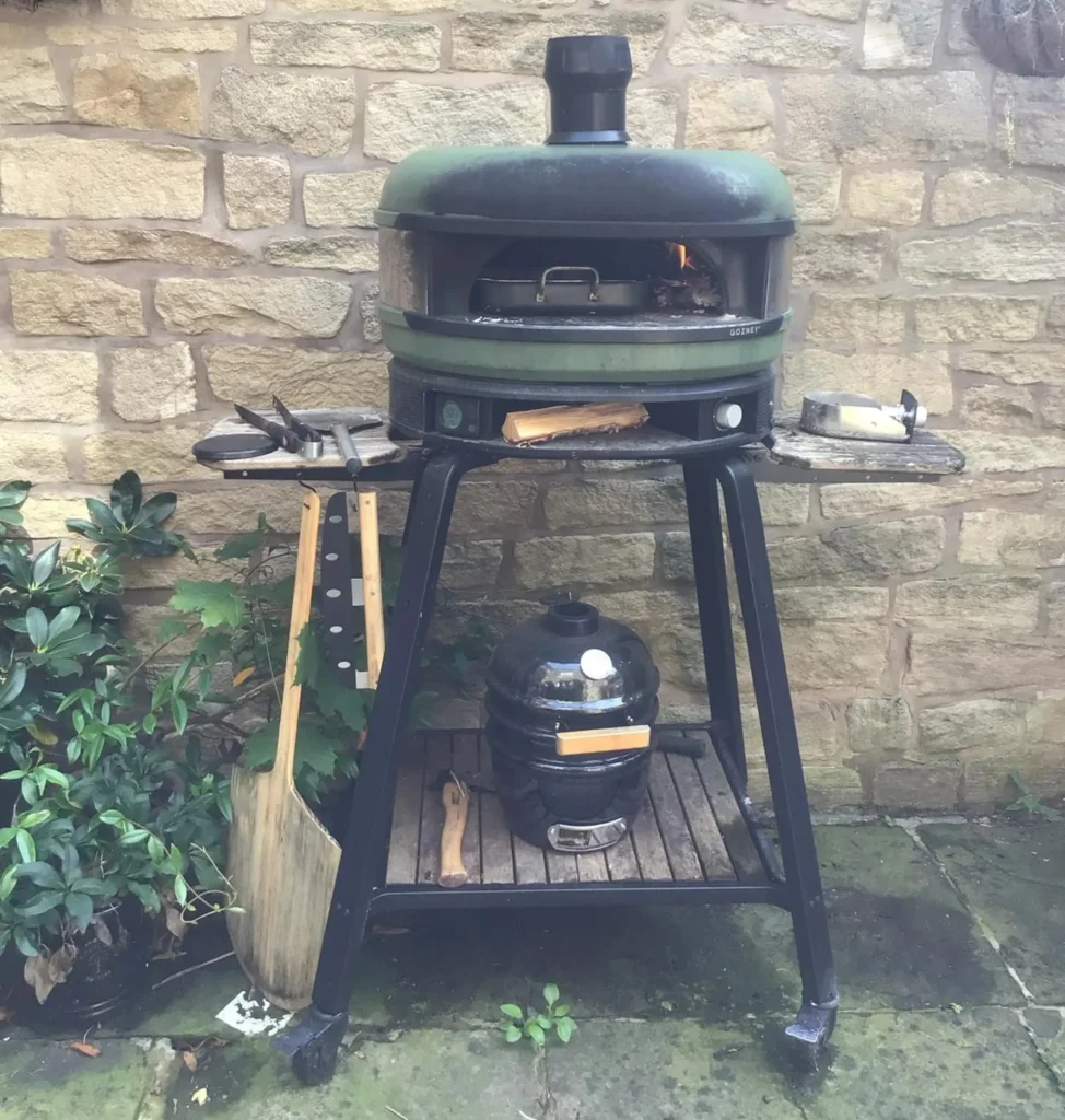 Gozney Dome on its stand in the Ramsbottom back garden — Lancashire stone wall, fairy lights, kamado smoker underneath, wood-fired oven at temperature
