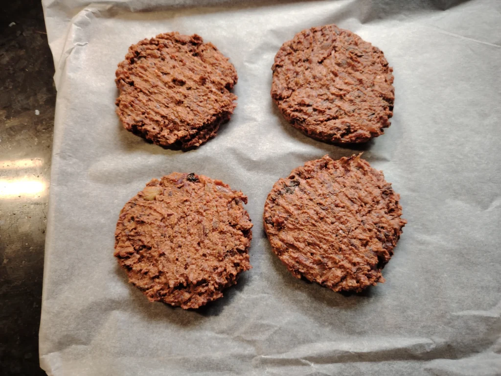 Four beetroot and black bean burger patties on baking parchment, black speckled granite worktop, ready to fry
