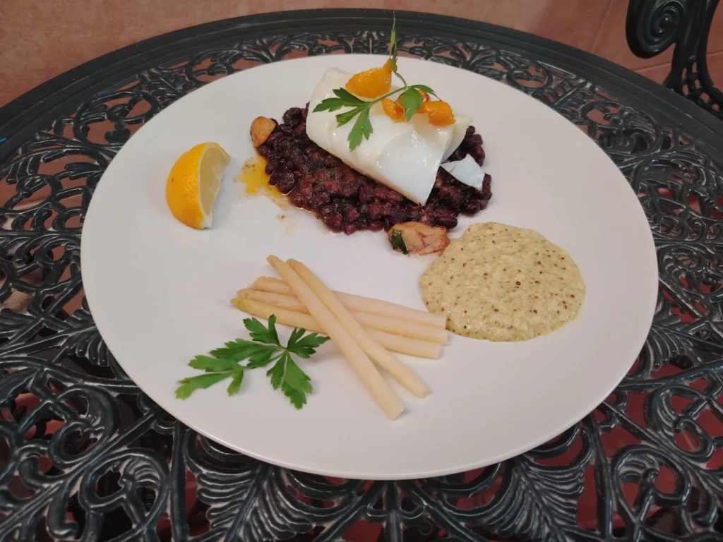Confit garlic recipe — olive oil poached cod loin on black beans with white asparagus, remoulade, confit lemon peel and parsley on a white plate on the Casa Adora patio