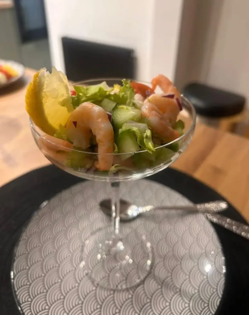 Prawn cocktail recipe — king prawns on shredded iceberg, cucumber and tomato with Rose Marie dressing in a coupe glass on a black plate, Rochdale