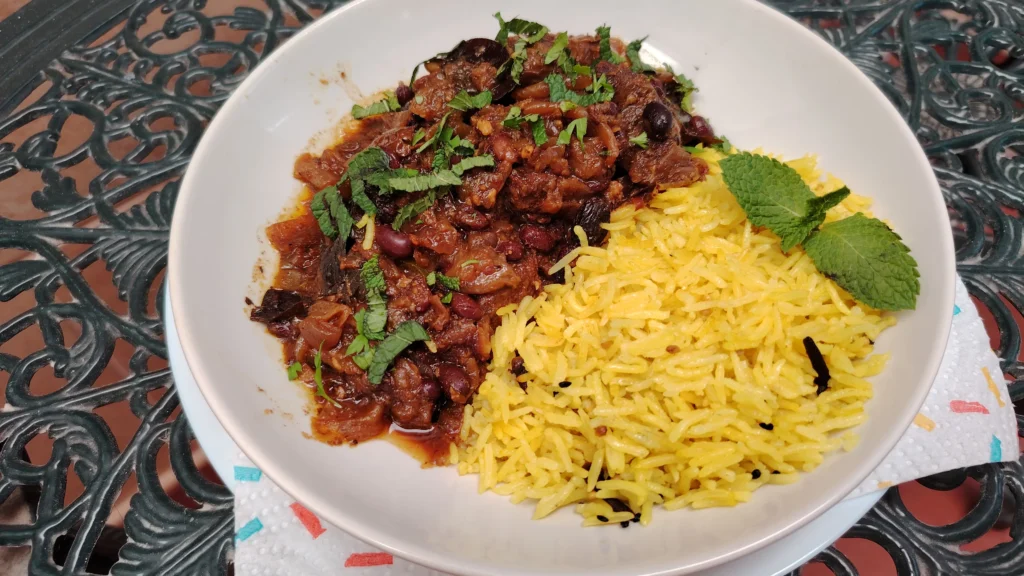 Kerala-style slow cooker beef curry with black beans in a white bowl, garnished with mint, served with turmeric basmati rice