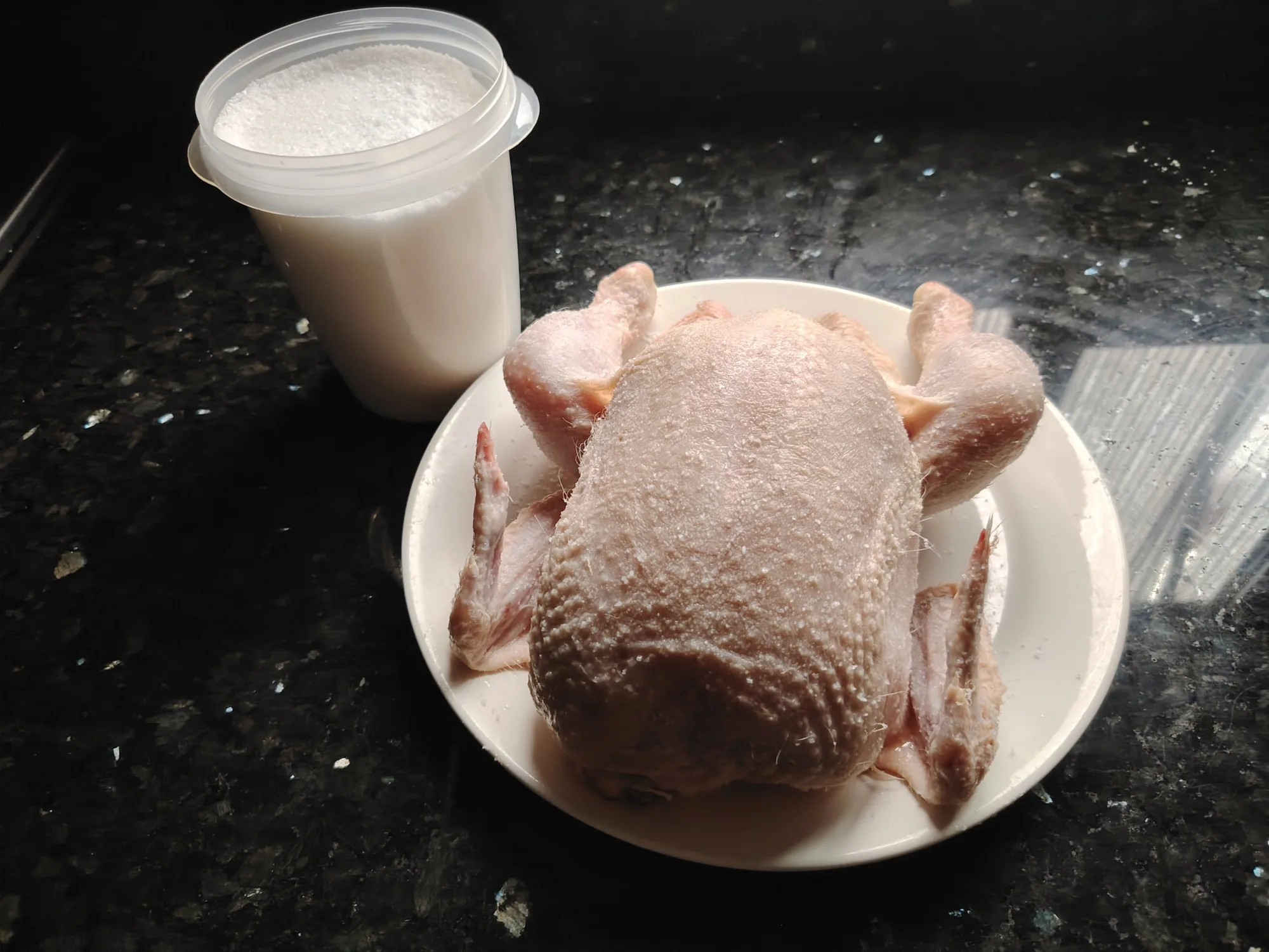 Raw whole chicken on a plate with dry, salted skin after dry brining, beside a tub of fine sea salt